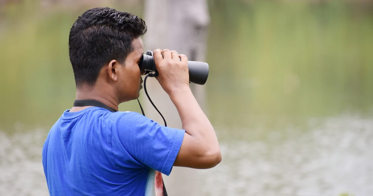 Homem observando aves à beira de um lago através de um binóculo